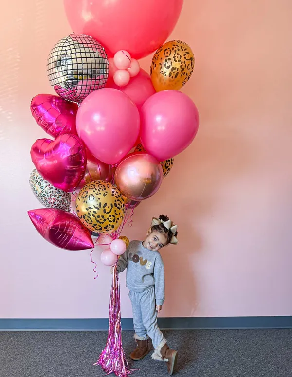 a woman standing next to a giant bunch of pink, gold, and leopard print balloons on a pink wall