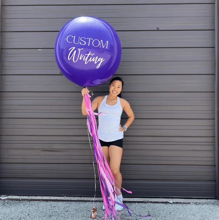 A woman is holding a purple balloon with the words "Happy Birthday" written on it, standing in front of a wall.