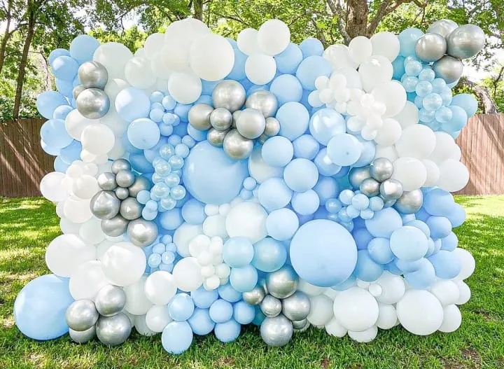 a bunch of blue and white balloons in the grass with a tree in the backgrouf of the photo