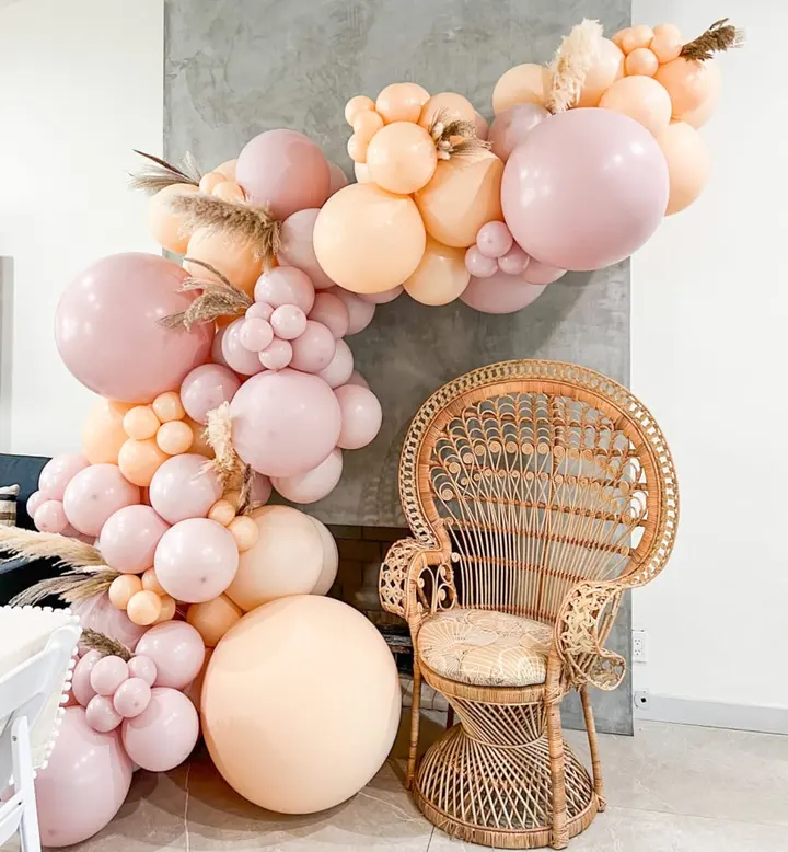 A vibrant arrangement of pink, peach, and white balloons adorns the foreground, complemented by a wicker chair in the background, creating a visually striking and whimsical scene.