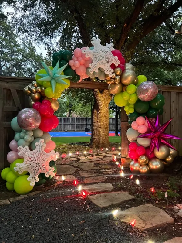 The image depicts a whimsical and colorful balloon arch installation in a backyard setting, with a tree in the background and a stone pathway leading towards the display.