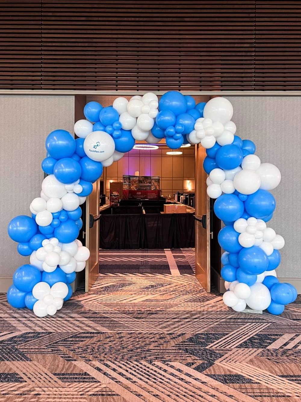 The image shows a decorative archway made of blue and white balloons, leading into a room with a dark interior and a carpeted floor.