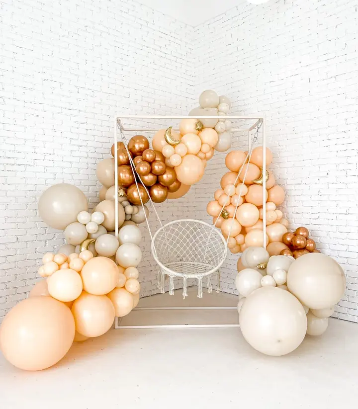 A wire frame chair surrounded by an assortment of balloons in various shades of beige, cream, and gold, set against a plain white background.