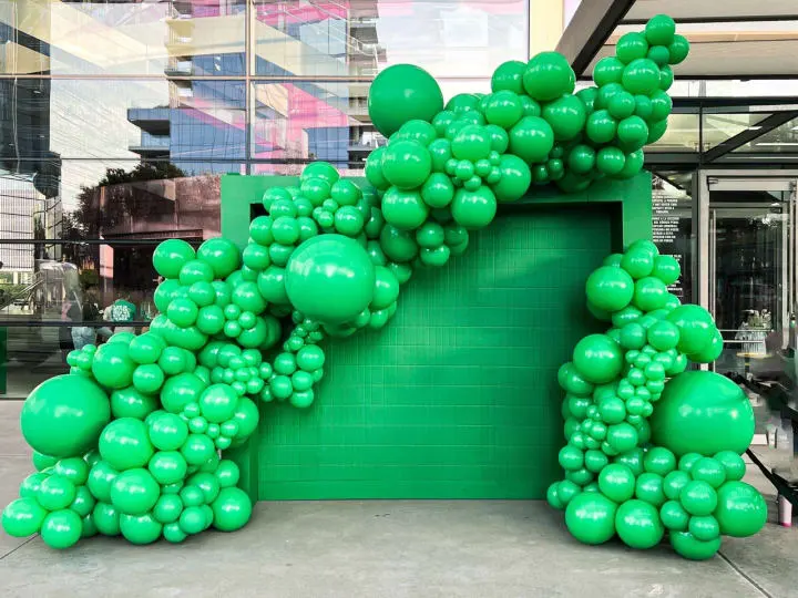 a large number of green balloons in front of a green wall with a green building in the back ground