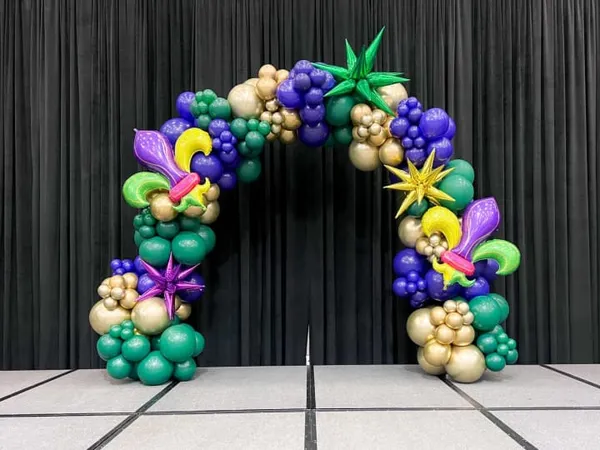 The image depicts a vibrant and colorful balloon arch adorned with a variety of balloon shapes and sizes, including grapes, stars, and flowers, set against a dark curtain backdrop.