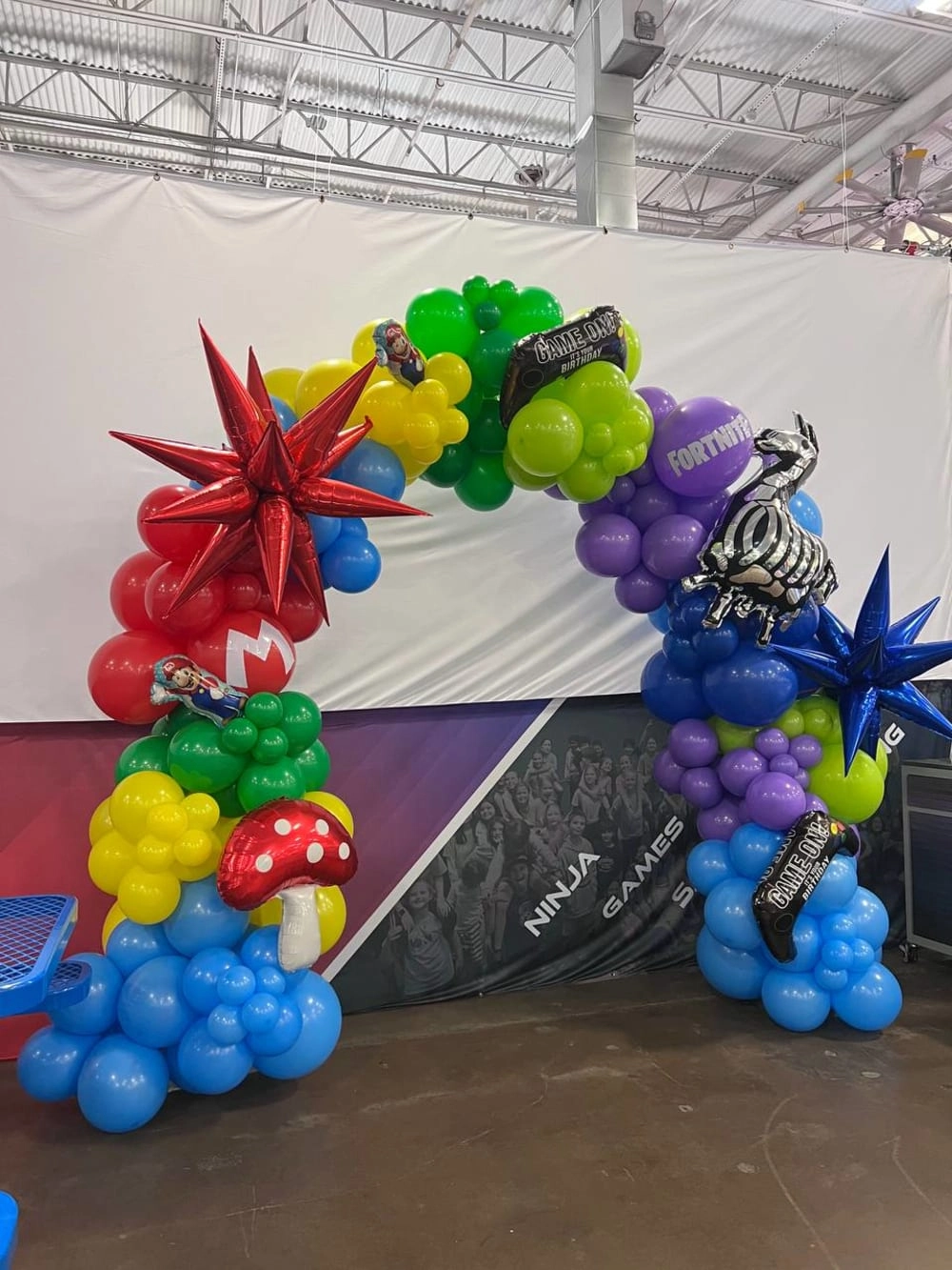 The image shows a colorful and whimsical balloon arch display featuring various shapes and designs, including mushrooms, stars, and other abstract forms, set against a plain white background in what appears to be an indoor event space or venue.