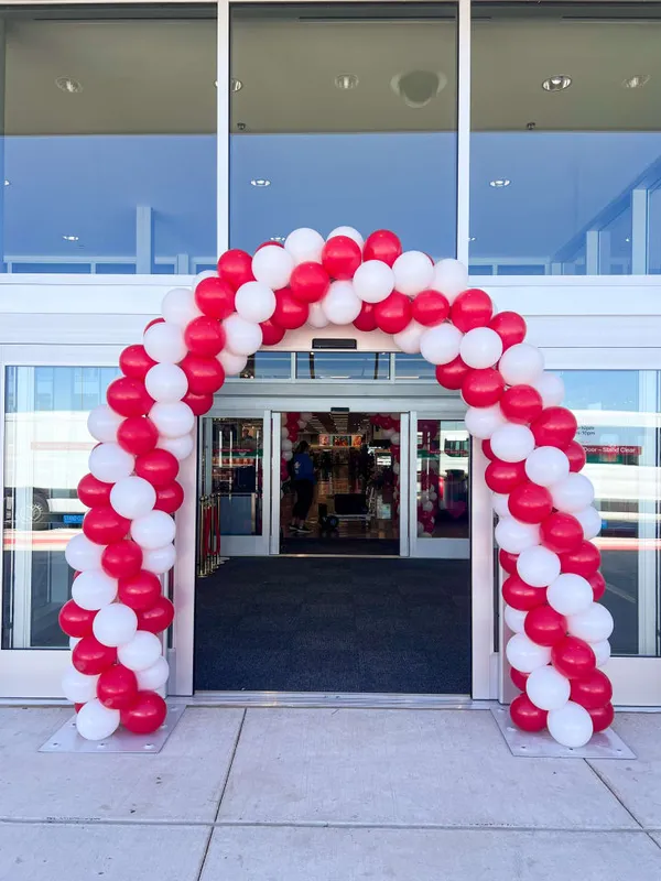 A large, colorful archway with red and white balloons is decorated with a red and white ribbon, creating a festive atmosphere. The archway is located in front of a building, and there are several people in the scene, indicating that it might be a special event or celebration.