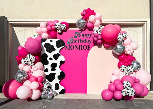 The image features a pink and black cow decorated with pink and black balloons, placed in front of a door.