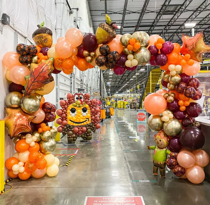 A vibrant and whimsical display of colorful balloons, autumn-themed decorations, and a smiling face sculpture, all set against a large industrial warehouse-like space.