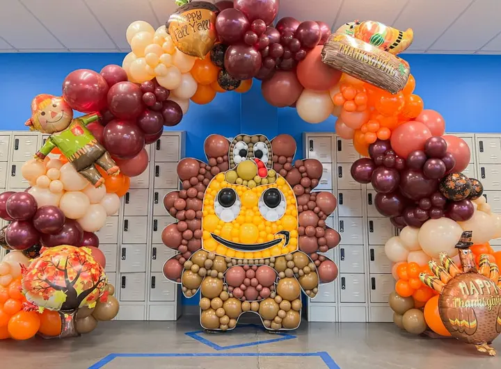 A vibrant and whimsical balloon arch made of various fruits and vegetables, including grapes, oranges, and pumpkins, stands in front of a row of lockers in what appears to be a school or office setting.