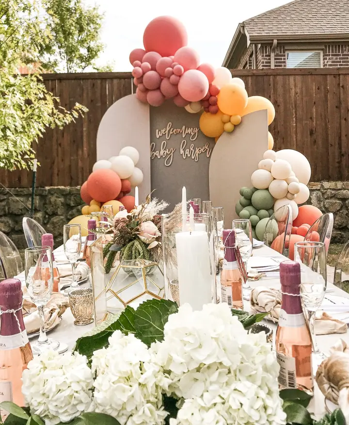 A beautifully decorated outdoor table setting with an array of colorful balloons, floral arrangements, and various decorative elements, all set against a backdrop of a wooden fence and lush greenery.