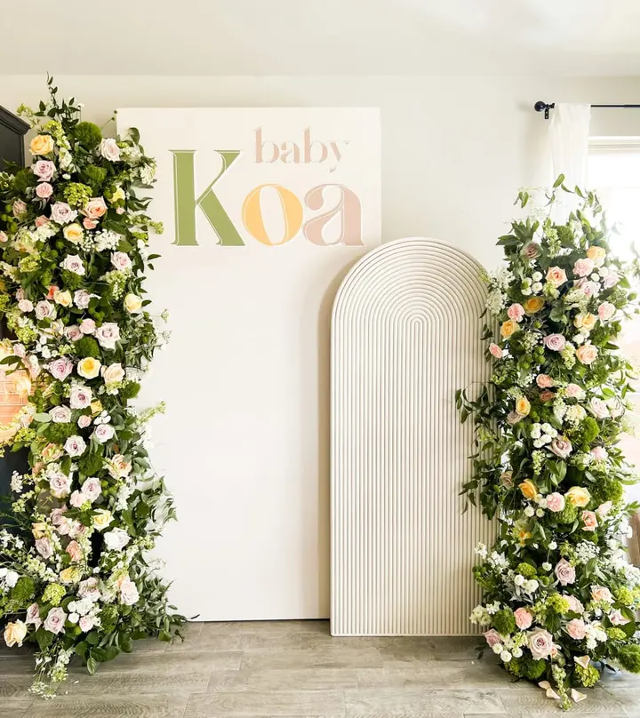 A floral archway adorned with lush greenery and vibrant flowers frames a sign that reads "Baby Koa", set against a neutral-toned background.