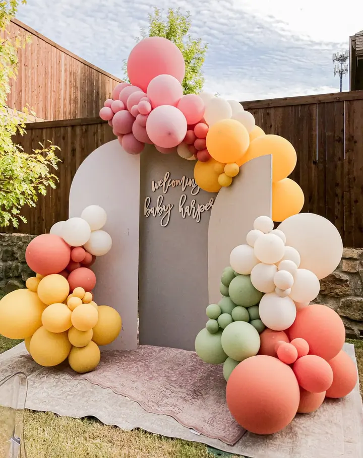 A colorful and whimsical balloon arch adorns the entrance to a baby shower event, with a variety of pastel-colored balloons of different sizes creating a vibrant and festive display against a wooden fence backdrop.