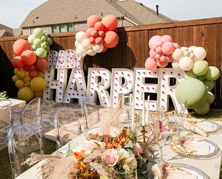 A festive outdoor setting with a large "HARPER" sign made of letters, surrounded by colorful balloons in various shades of pink, red, and white, and a table decorated with floral arrangements and place settings.