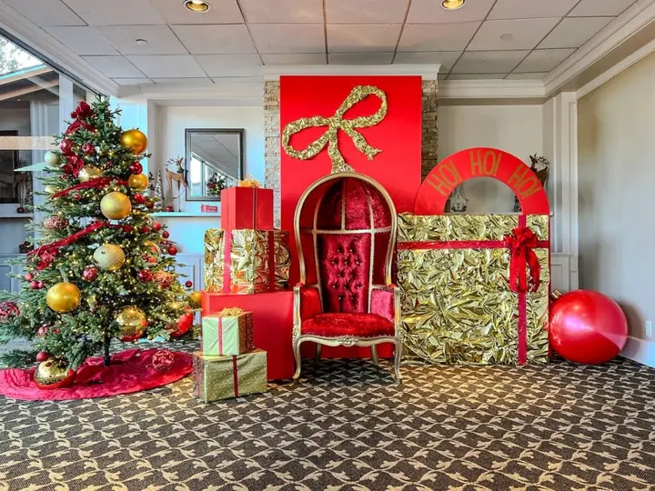 A festively decorated room with a Christmas tree, ornaments, and a large red and gold throne-like chair surrounded by wrapped gifts and other holiday decor.