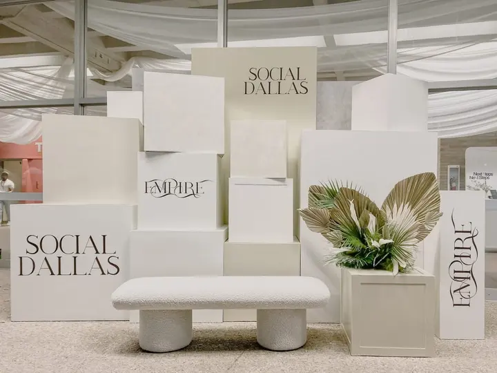 The image depicts a minimalist and modern display setup, featuring white podiums and pedestals with the text "Social Dallas" prominently displayed. The background is adorned with draped fabric, creating a visually appealing and cohesive aesthetic.