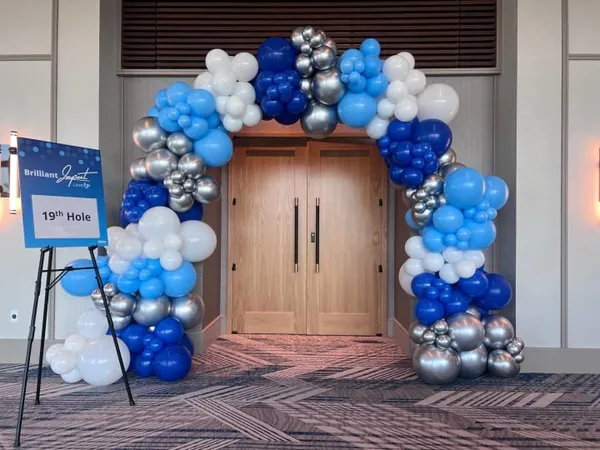 The image shows a decorative balloon arch in shades of blue, white, and silver, framing the entrance to a building. The balloons are arranged in a whimsical and festive manner, creating a welcoming and celebratory atmosphere.