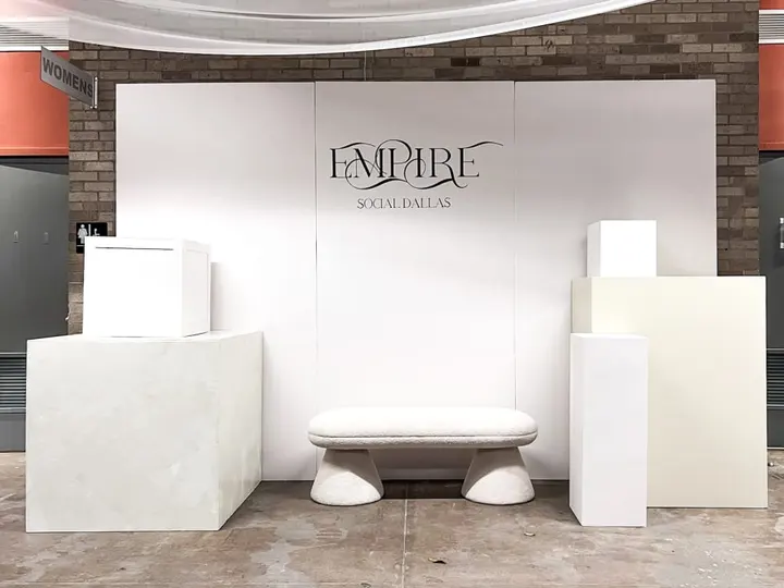 A minimalist display setup with a large white backdrop featuring the "Empire" logo, surrounded by various white pedestals and platforms on a tiled floor.