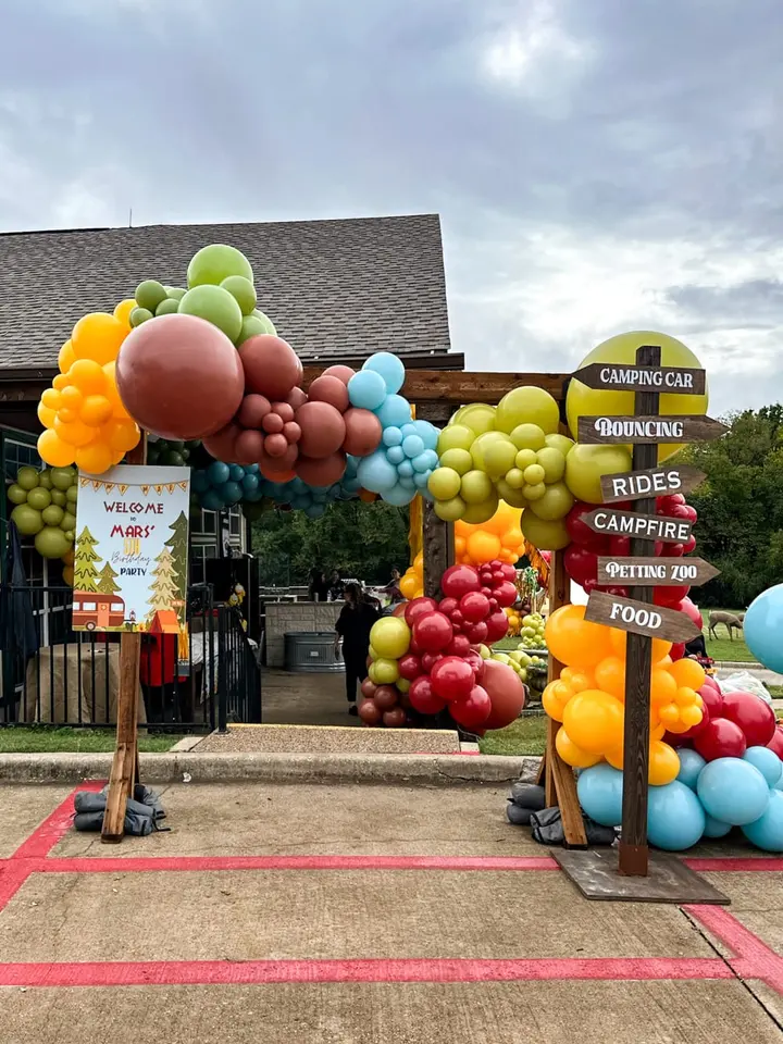 A colorful display of large inflatable balloons in various shapes and sizes, including fruits and other objects, stands in the foreground, while a wooden building with signage for a camping and recreational vehicle business can be seen in the background.