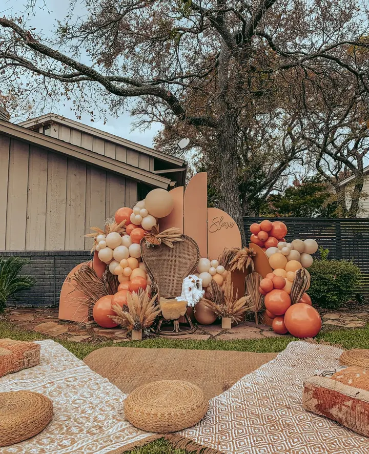 A vibrant autumn display featuring an assortment of pumpkins, gourds, and other seasonal decorations arranged in the foreground, set against a backdrop of a wooden building and a lush, tree-lined landscape.