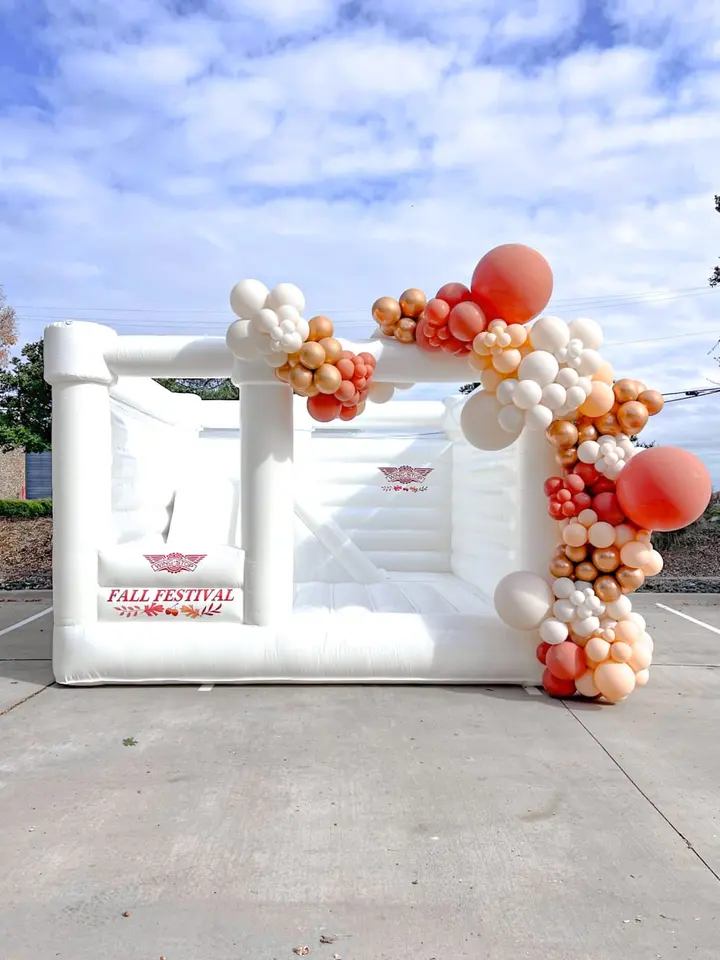 A large white inflatable structure with colorful balloon clusters decorating the exterior, set against a blue sky with scattered clouds in an outdoor setting.