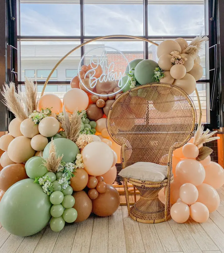 A vibrant and whimsical display of pastel-colored balloons, greenery, and a wicker chair set against a large window, creating a warm and inviting atmosphere.