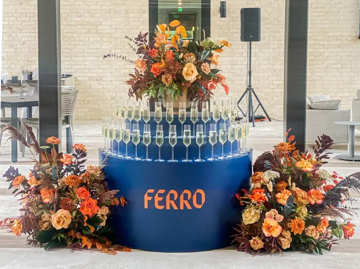 A large, ornate floral arrangement sits atop a blue platform with the word "FERRO" prominently displayed, surrounded by a display of wine glasses in a modern, industrial-style setting.