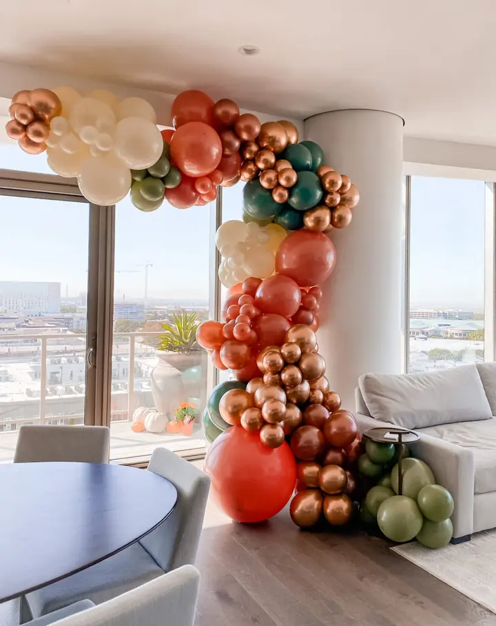 A colorful and whimsical balloon arch made of various shades of orange, red, and green balloons dominates the foreground, while a panoramic view of a city skyline can be seen through the large windows in the background.