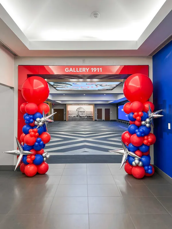 The image shows a decorative balloon arch in red and blue colors, framing the entrance to a gallery or event space called "Gallery 1911". The background features a tiled floor and a large display or screen on the wall.