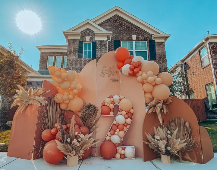 A large, colorful display of autumn-themed decorations, including balloons, pumpkins, and dried foliage, stands in the foreground, with a brick house visible in the background.