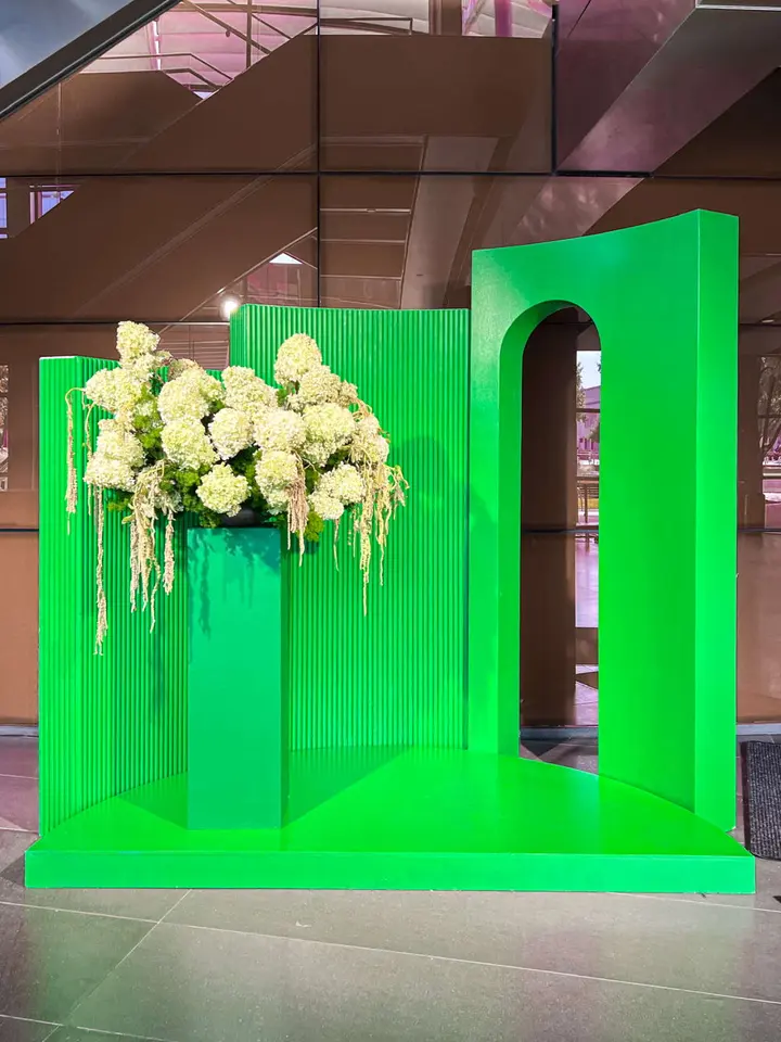 The image depicts a vibrant green display with large white floral arrangements, set against a backdrop of brown architectural elements.