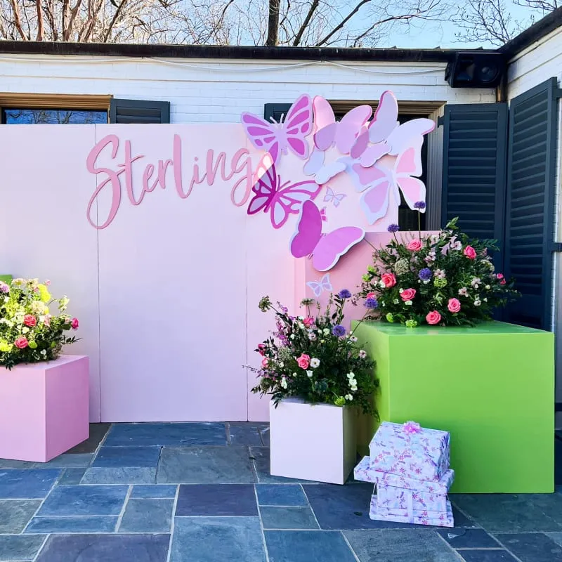 The image shows a decorative backdrop with the name "Sterling" written in cursive, surrounded by large pink and purple flowers. In the foreground, there are several potted plants and floral arrangements on green and pink pedestals, creating a vibrant and festive display.