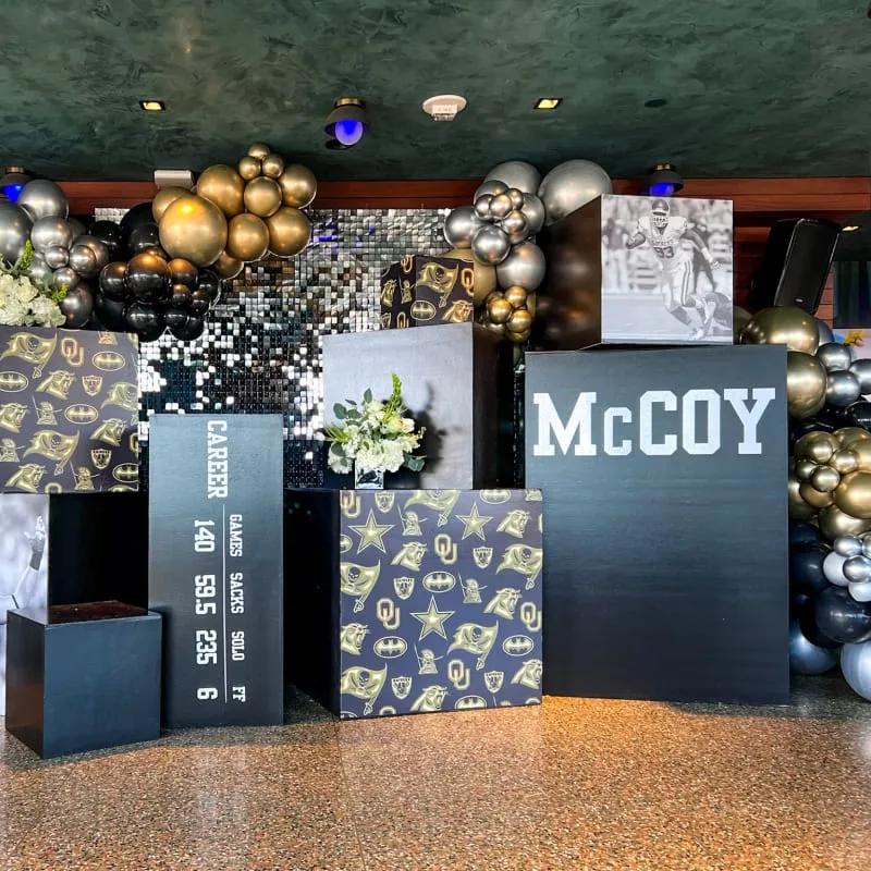 The image shows an elaborate display of gold and black balloons, along with various decorative elements, creating a festive and celebratory atmosphere. The focal point appears to be the "McCoy" signage, suggesting this is likely a celebration or event related to that name or brand.