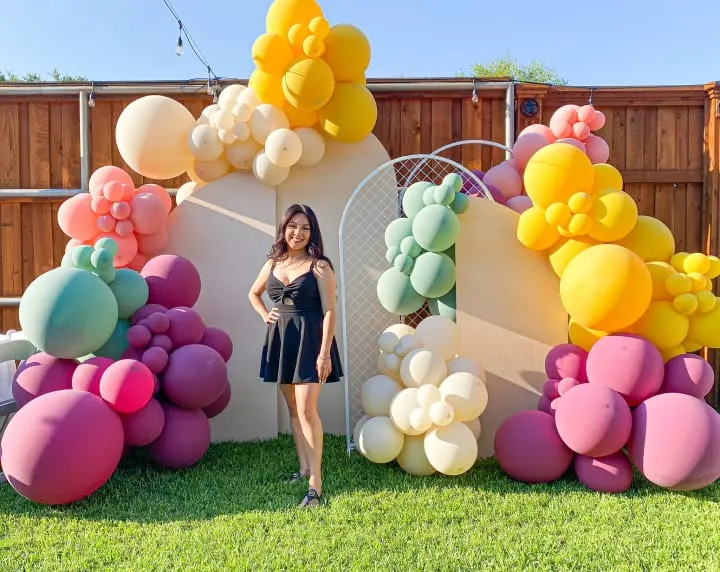 a woman standing in front of a bunch of balloons on a fenced in area with grass and a fenced in area behind her