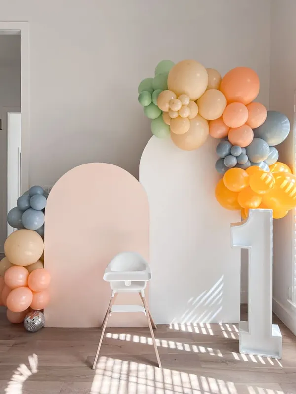 The image features a room with a white wall and a large, colorful balloon archway. The room is decorated with balloons, including a large one in the center and smaller ones scattered around. There are also two chairs placed in the room, one on the left side and another on the right side.