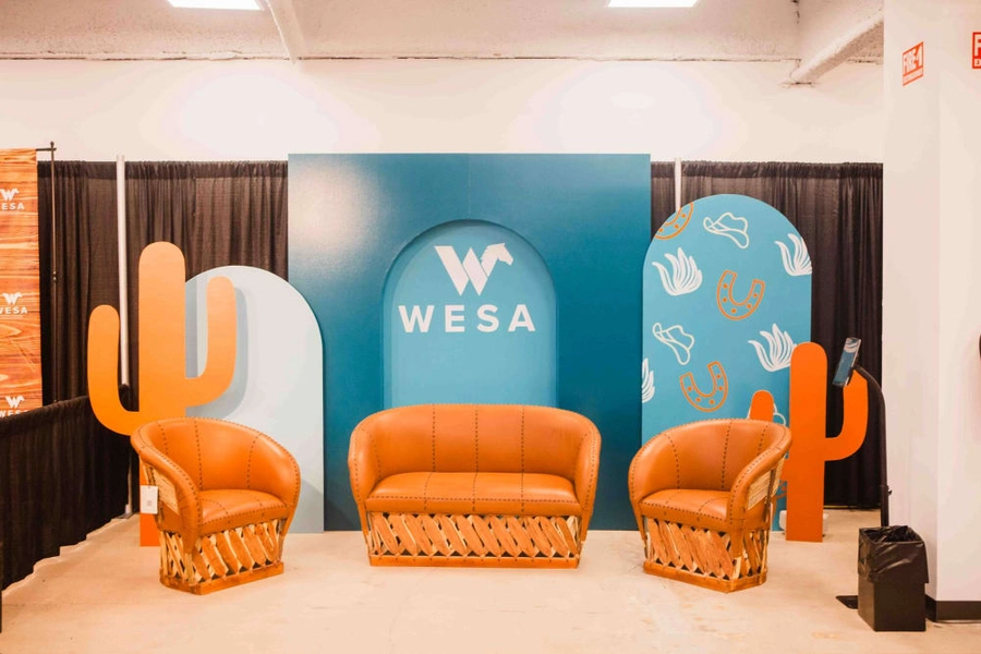 The image depicts a modern and vibrant office or event space, featuring a large blue backdrop with the "WESA" logo, surrounded by orange wicker chairs and decorative cactus-like shapes on the walls.