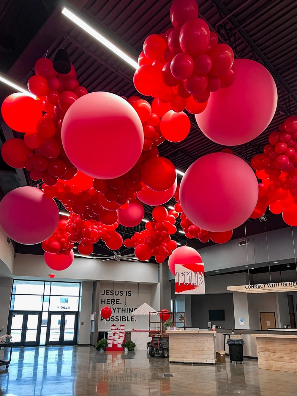 The image shows a large indoor space with a ceiling covered in numerous red balloons of various sizes, creating a vibrant and festive atmosphere.