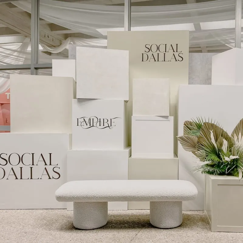 The image shows a minimalist display with white rectangular structures, a potted plant, and signage for "Social Dallas" in the foreground, set against a backdrop of draped fabric.