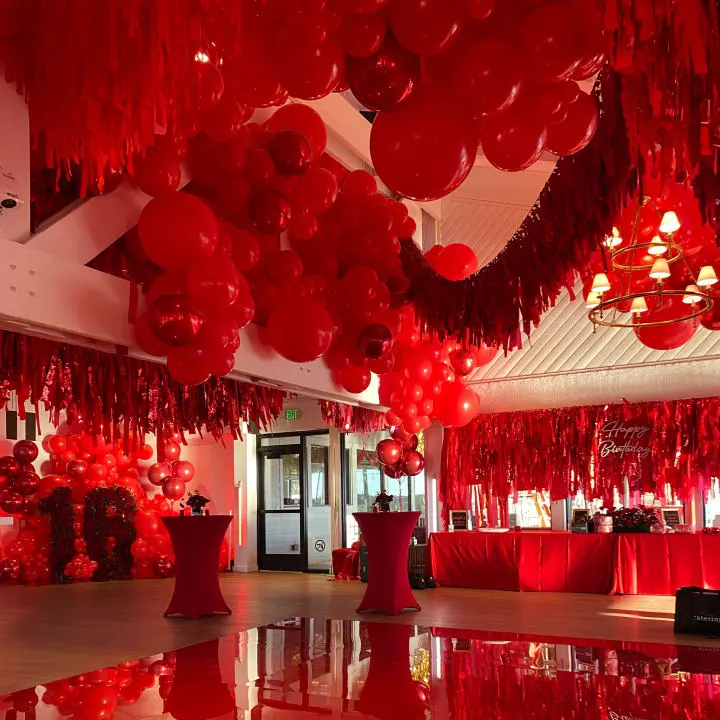a room filled with lots of red balloons hanging from the ceiling and a table with red cloths on it