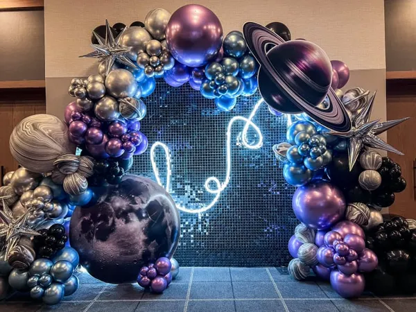 The image depicts a whimsical and vibrant space-themed balloon arch, featuring an array of colorful balloons in various shapes and sizes, including planets, stars, and abstract shapes, set against a backdrop of a glittering, mosaic-like surface.