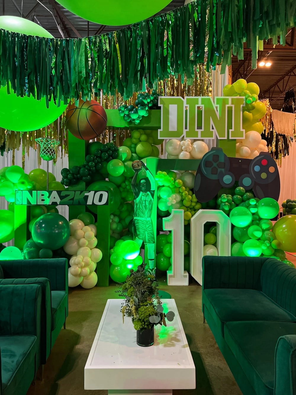 A vibrant and festive green-themed party setup, featuring various decorative elements such as hanging streamers, balloons, and a large "DINI" sign, along with gaming-related props like a controller, creating an immersive and engaging atmosphere.