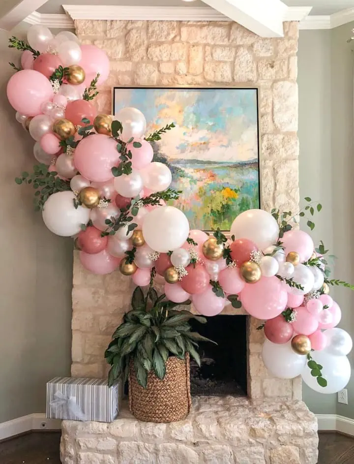 a fireplace decorated with pink, white, and gold balloons and a potted plant in front of it