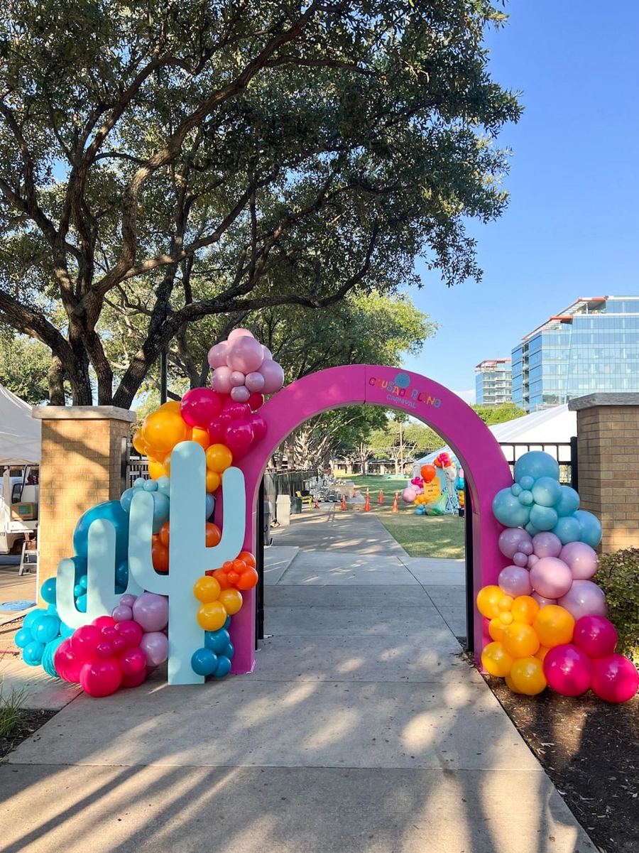 A colorful balloon arch adorns the entrance to an outdoor event, surrounded by lush trees and a clear blue sky in the background.