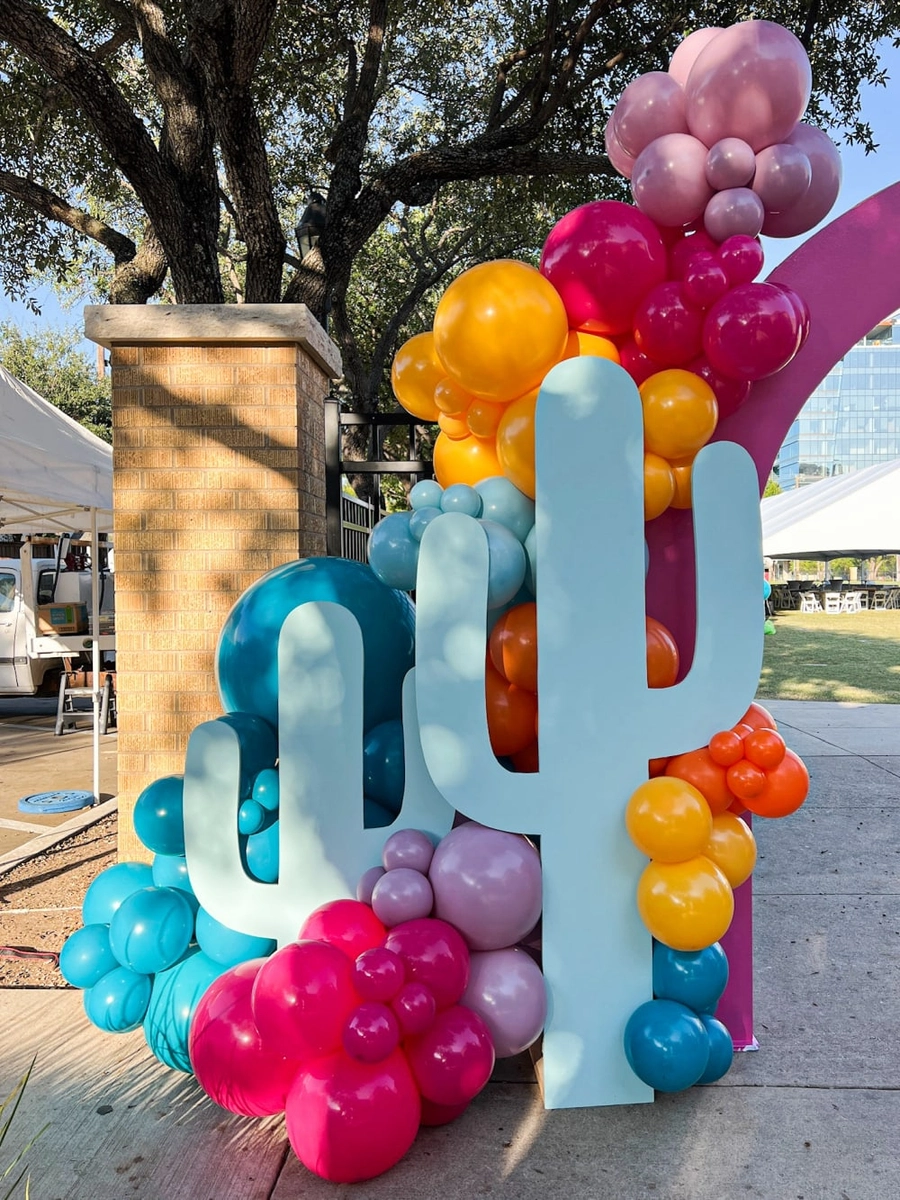 Colorful balloon sculptures in the shape of cacti and other shapes are displayed in an outdoor setting with a large oak tree in the background.