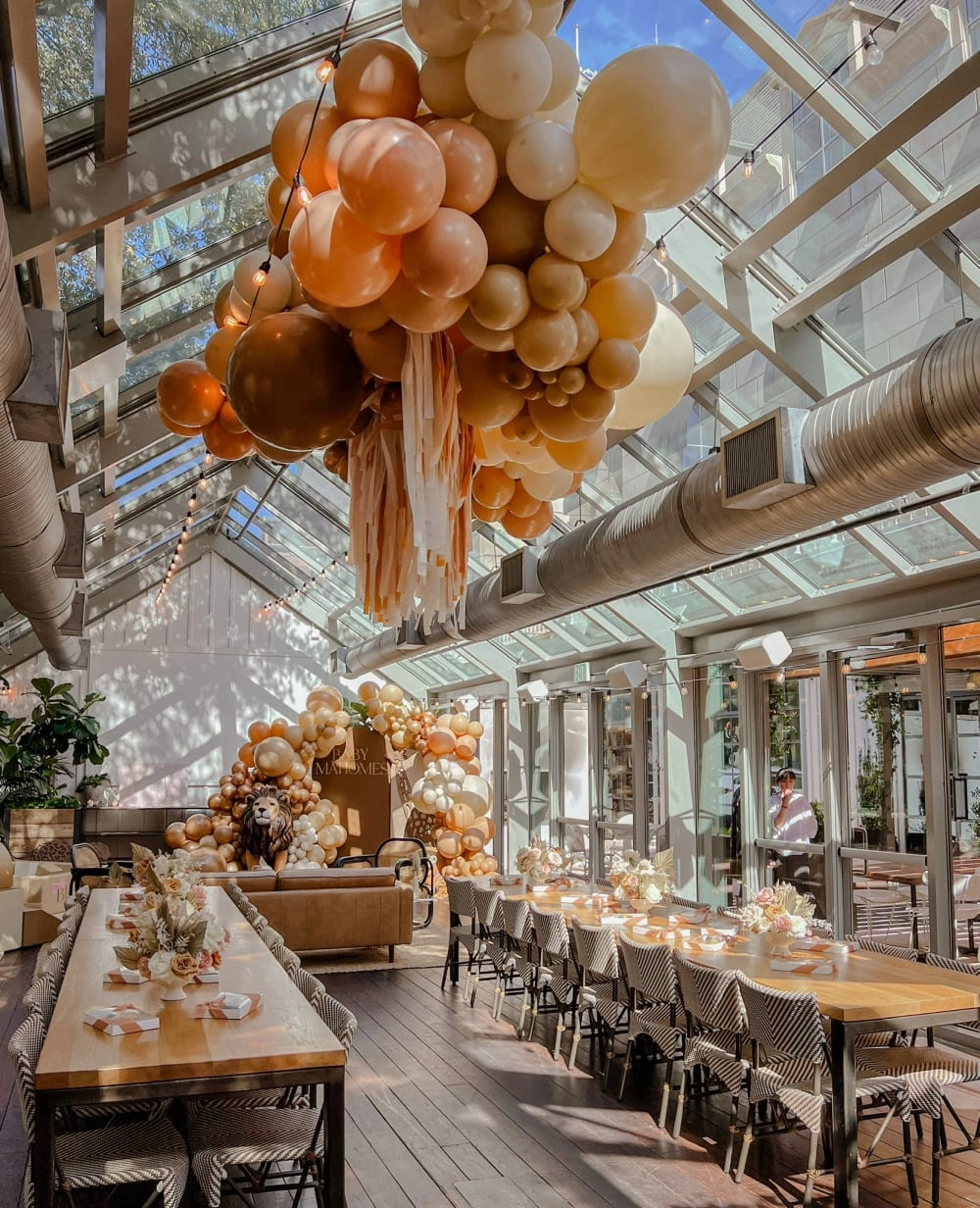 A large, open-plan indoor space with a high ceiling, featuring an elaborate arrangement of colorful balloons in various shades of orange and yellow suspended from the ceiling, creating a warm and festive atmosphere. The foreground shows a long wooden table with chairs, suggesting a dining or event setting.