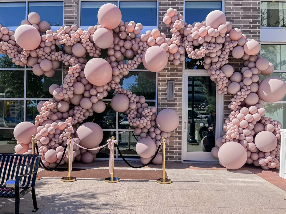 A large, whimsical balloon arch in shades of pink and white adorns the entrance of a building, creating a vibrant and eye-catching display against the backdrop of the brick structure.