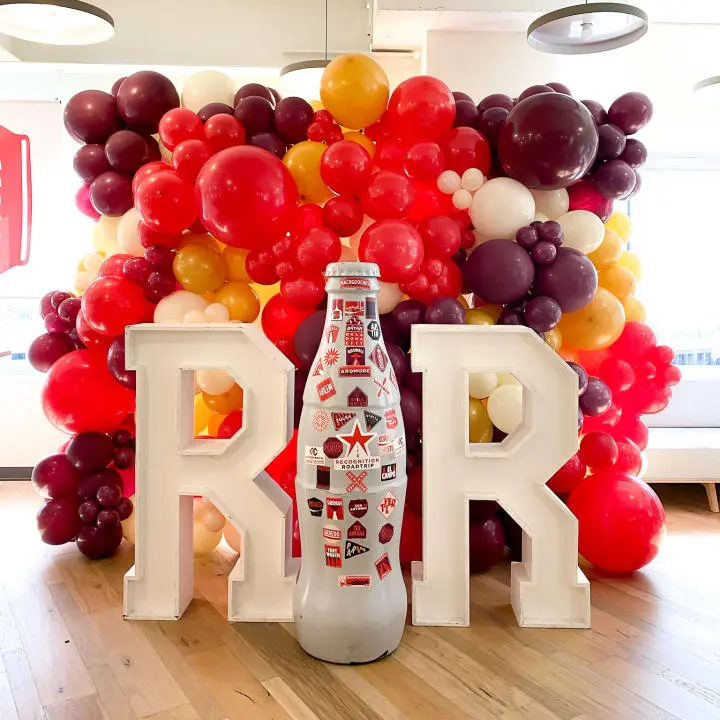 a bottle of soda sitting on top of a wooden floor in front of a large balloon wall with letters and balloons