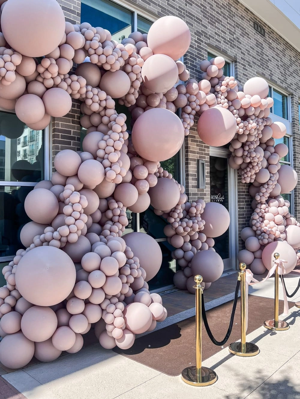 A large, whimsical display of pink and white balloons of various sizes cascades down the exterior of a brick building, creating a visually striking and playful scene.