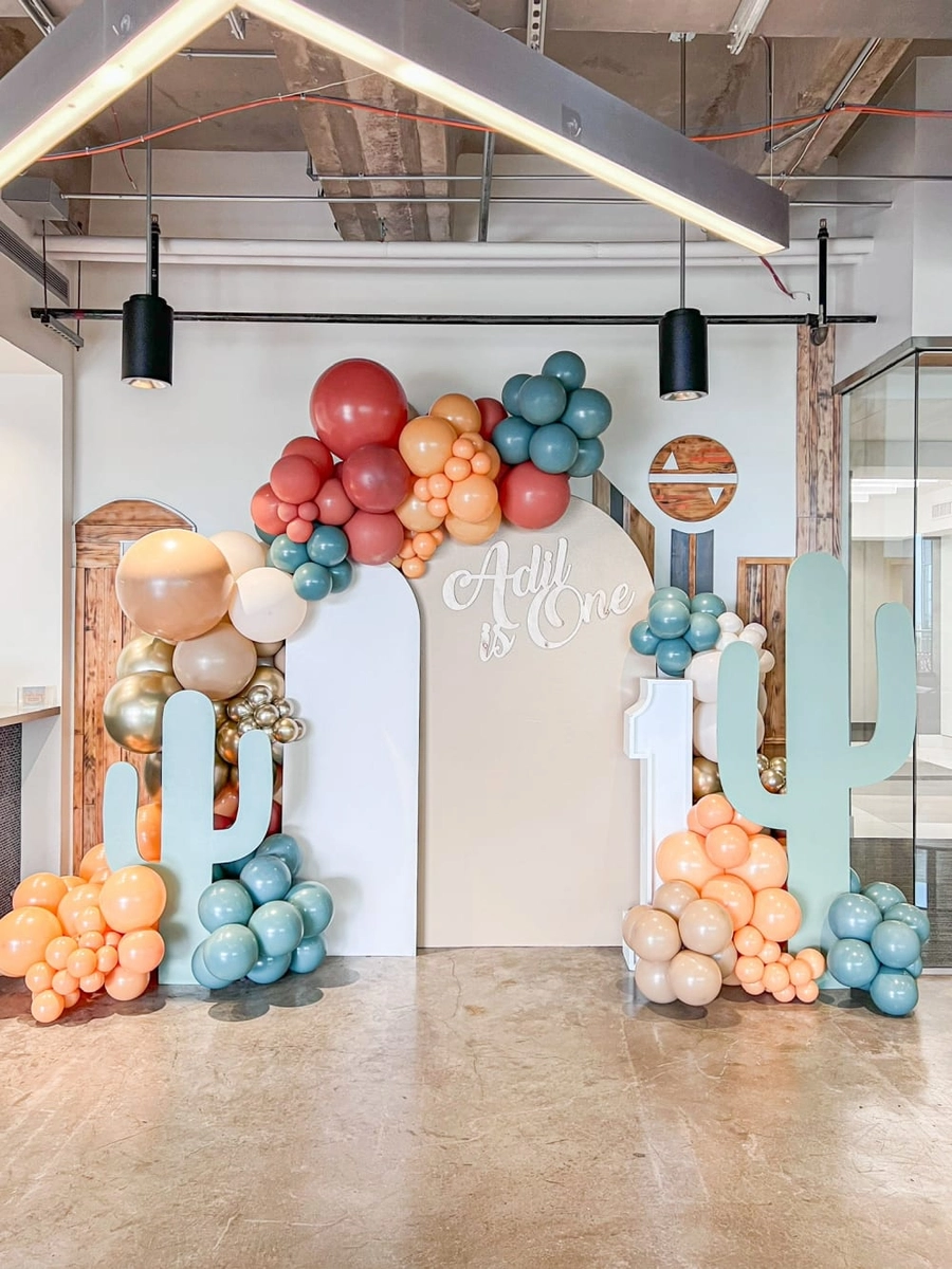 A colorful balloon arch with various shades of orange, blue, and white balloons decorates the entrance of a modern, industrial-style space with exposed beams and concrete floors.