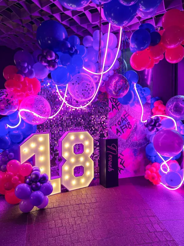 The image depicts a vibrant and colorful party or event setting, with large illuminated numbers "18" in the foreground surrounded by an array of balloons, neon lights, and other decorative elements in shades of purple, pink, and blue.